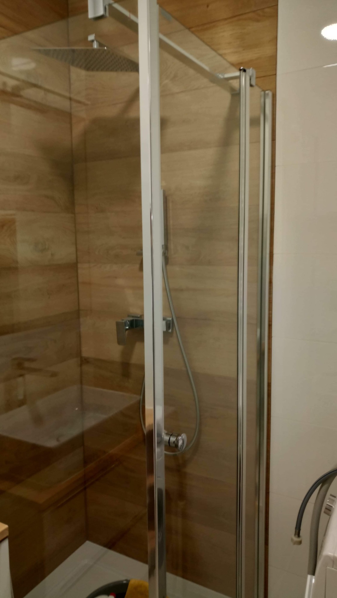 Szklana kabina prysznicowa z deszczownicą i baterią, wykończona płytkami imitującymi drewno na ścianach, widoczna częściowo pralka z szarym wężem odpływowym.