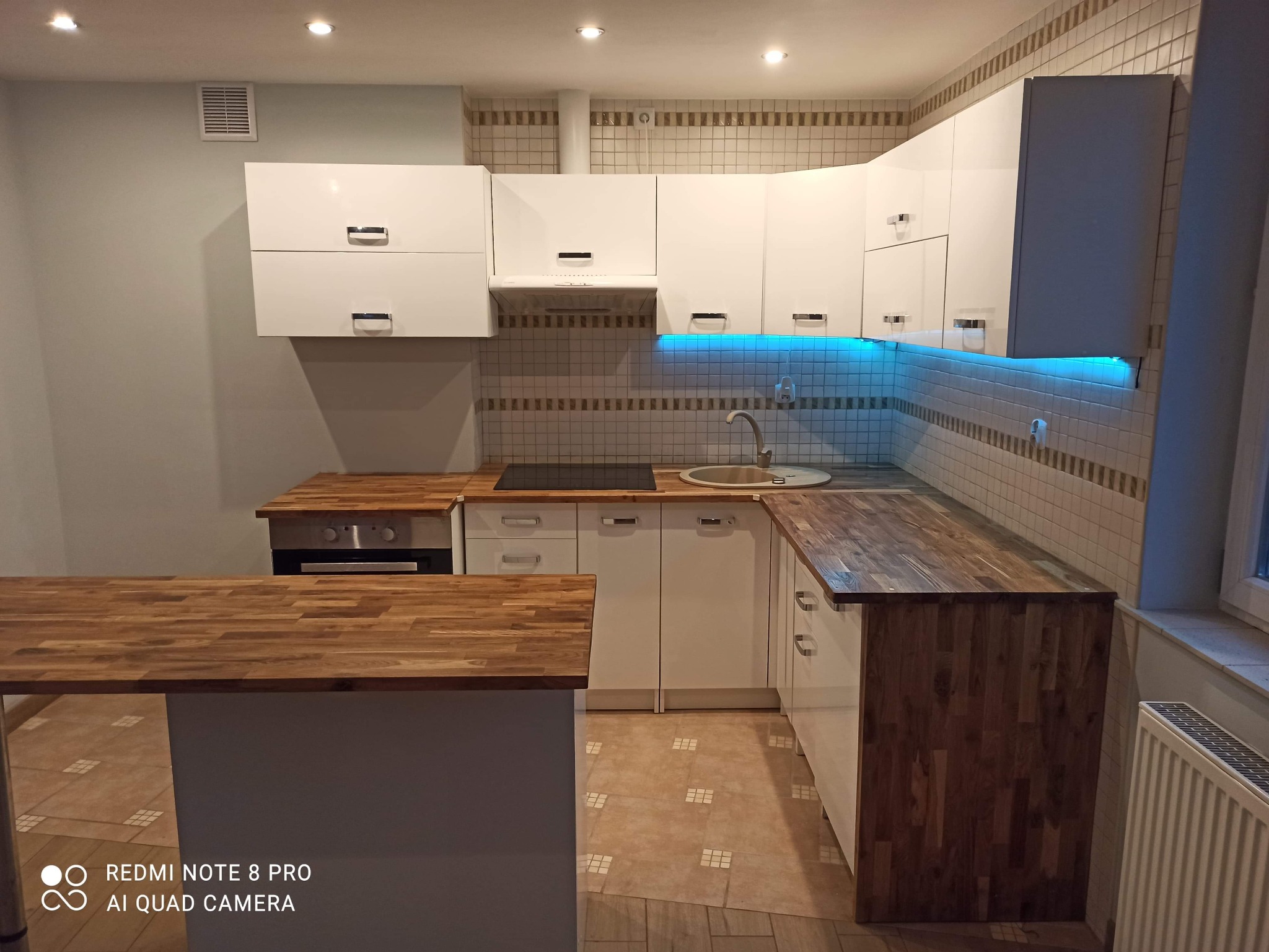 Nowoczesna kuchnia z białymi szafkami, drewnianym blatem i podświetleniem LED pod szafkami wiszącymi, widok na wyspę kuchenną.