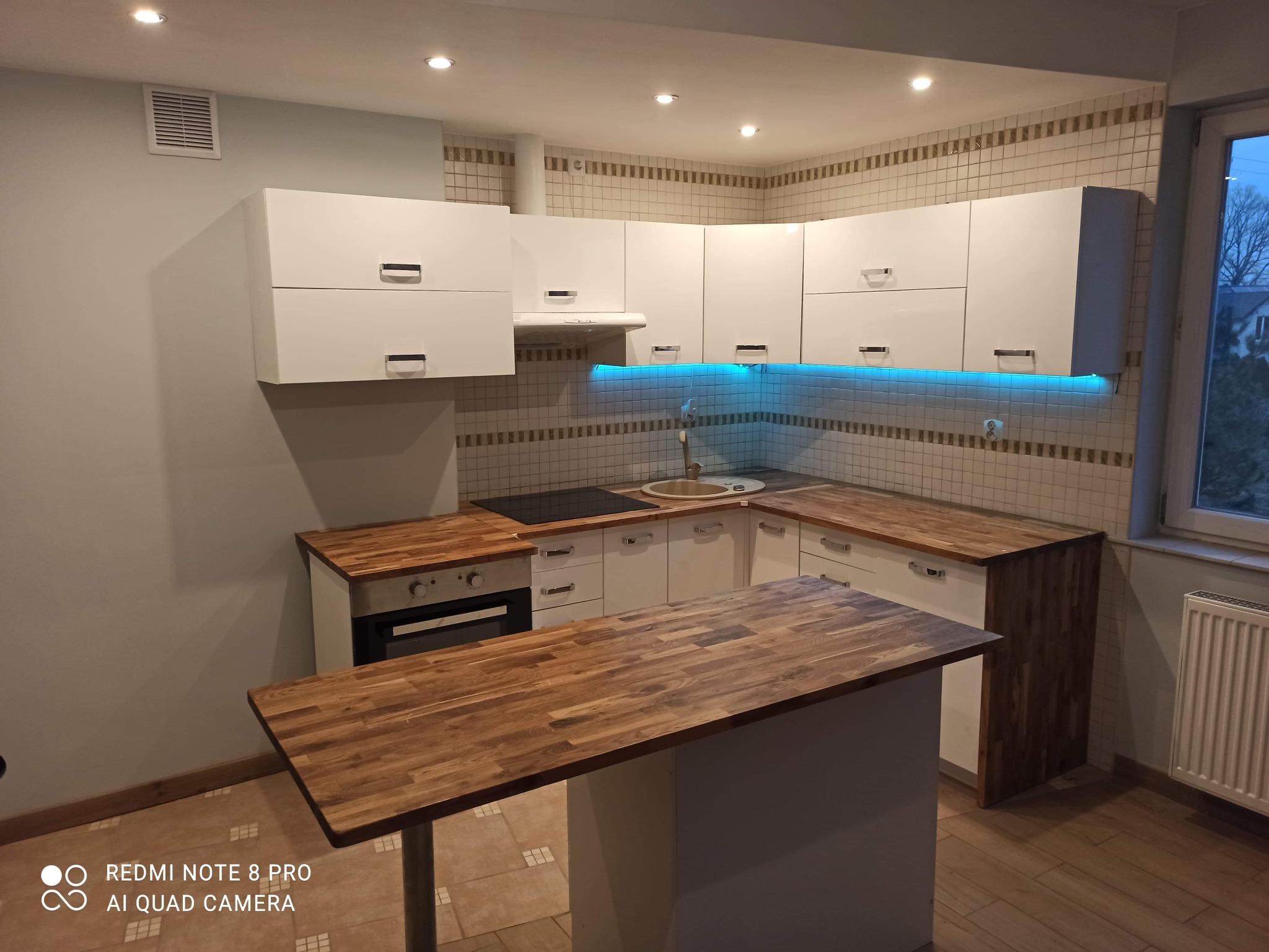 Nowoczesna kuchnia z białymi szafkami, drewnianymi blatami i podświetleniem LED pod szafkami wiszącymi, widoczna wyspa kuchenna z drewnianym blatem.