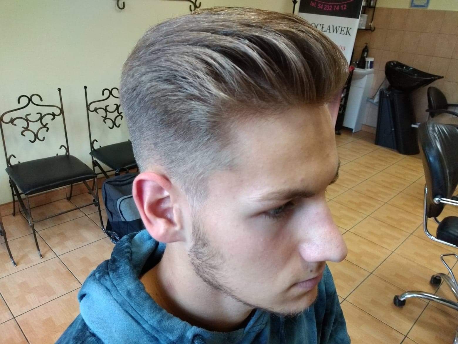 Męska fryzura z wycieniowanymi bokami i zaczesanymi do góry włosami, widok z boku w salonie fryzjerskim z krzesłami o metalowej konstrukcji w tle.