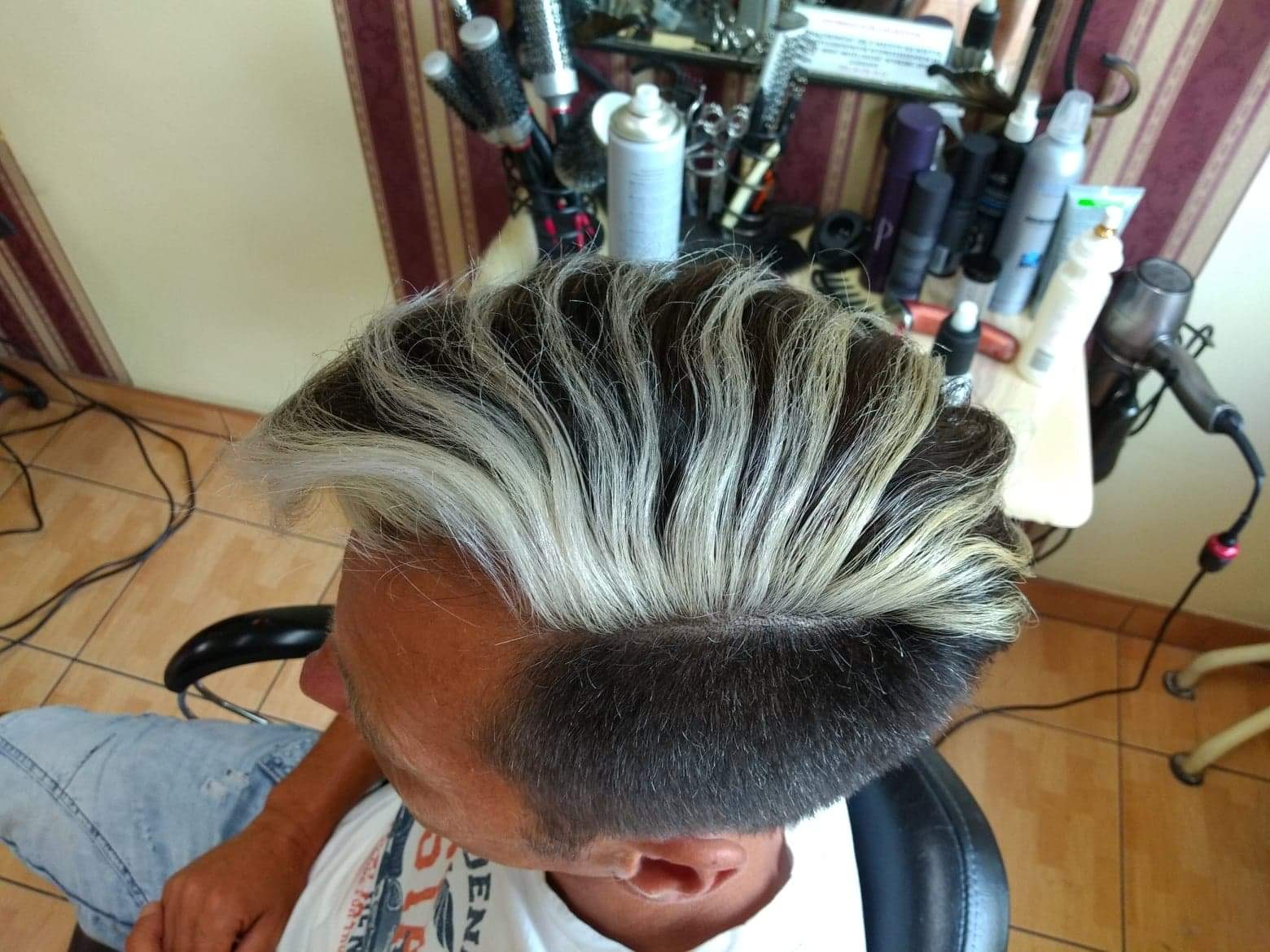 Męska fryzura z kontrastowym, platynowym pasem na górze, widok z góry, w tle półka z kosmetykami i akcesoriami.