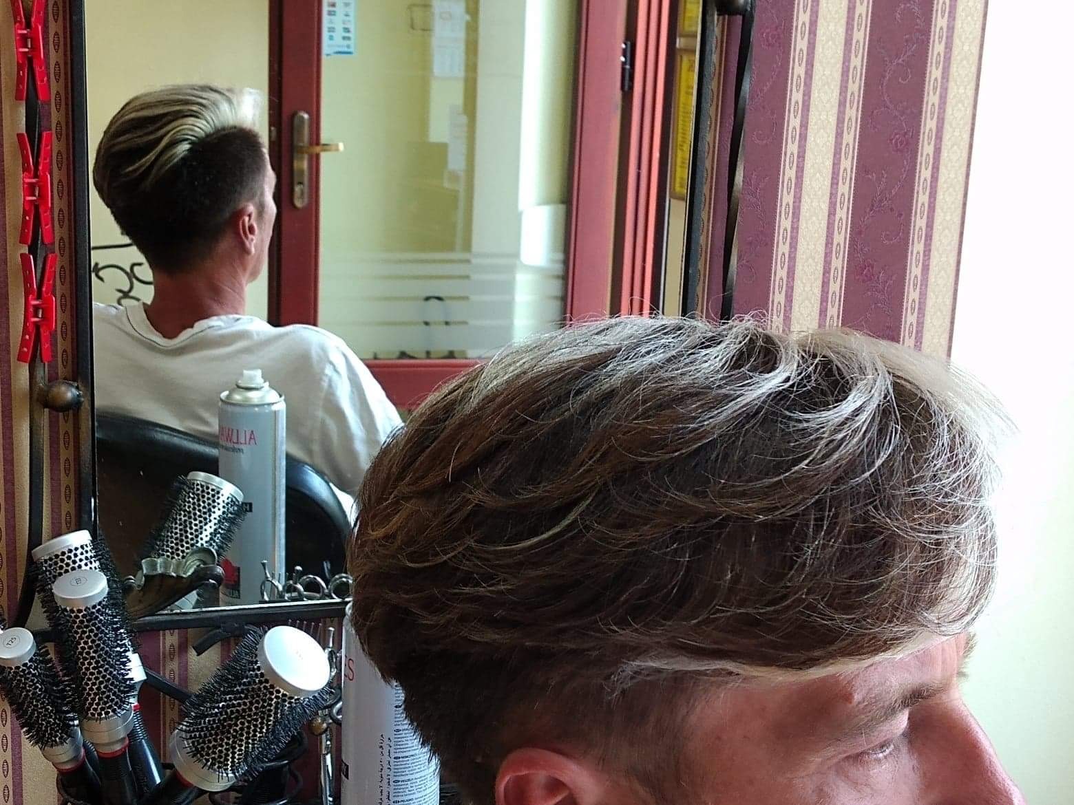 Widok z bliska na świeżo ostrzyżone włosy z jasnymi pasemkami, odbicie w lustrze ukazuje klienta w trakcie strzyżenia, a obok widoczne narzędzia i kosmetyki.