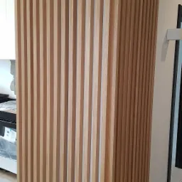 Drewniana ścianka działowa z pionowych listew, widok z boku, jasne drewno, częściowo zasłania wnętrze kuchni.