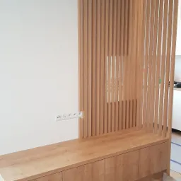 Drewniana zabudowa z szafkami i ażurową ścianką działową z pionowych listew, widoczne gniazdka elektryczne na ścianie, podłoga zabezpieczona kartonem.