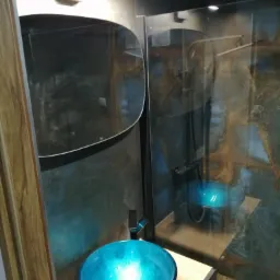 Błękitna szklana umywalka nablatowa z chromowaną baterią w nowoczesnej łazience z kabiną prysznicową typu walk-in i ścianami wykończonymi płytkami imitującymi kamień.