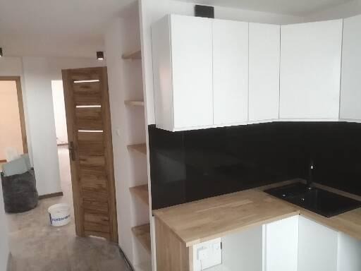 Częściowy widok nowocześnie wykończonej kuchni z białymi szafkami górnymi, czarnym panelem nad blatem, drewnianym blatem i widoczną umywalką; w tle drzwi z jasnego drewna oraz wiadro po farbie...