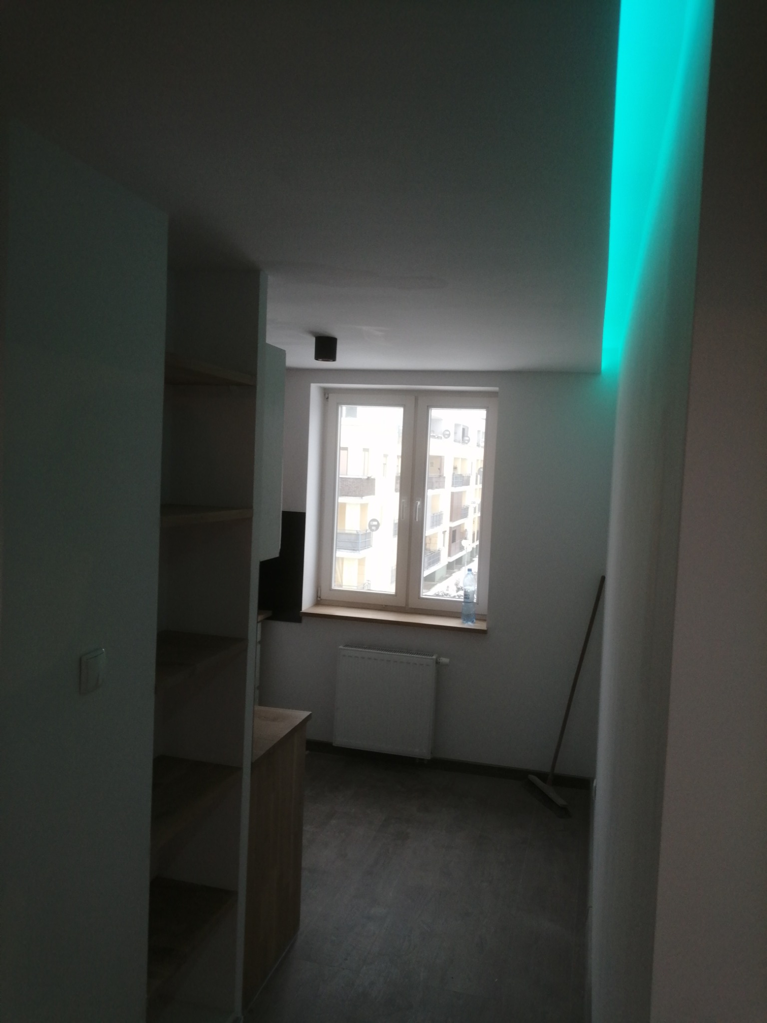 Wnętrze mieszkania po remoncie, widok na okno z grzejnikiem, fragment szafki z półkami i podświetlany na zielono sufit.