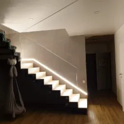 Nowoczesne schody z podświetlanymi stopniami i szklaną balustradą, widok z dołu, w tle drewniana podłoga i szara ściana.