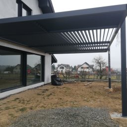 SPAW-TEAM Tomasz Ziebura - Nowoczesna, grafitowa altana z lamelowym dachem, przylegająca do budynku z dużymi oknami. Widoczne słupy i przygotowany teren pod dalsze prace.