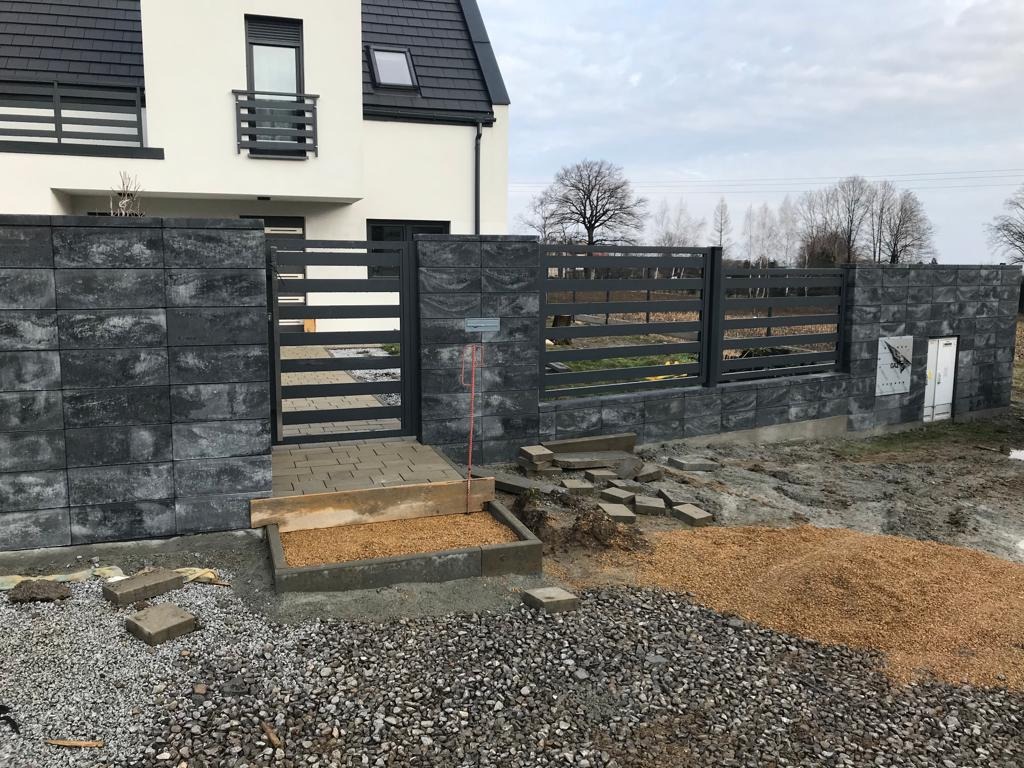 Nowoczesne ogrodzenie z szarej kostki łupanej i metalowych przęseł, z furtką i schodami wejściowymi, w trakcie prac wykończeniowych na posesji.