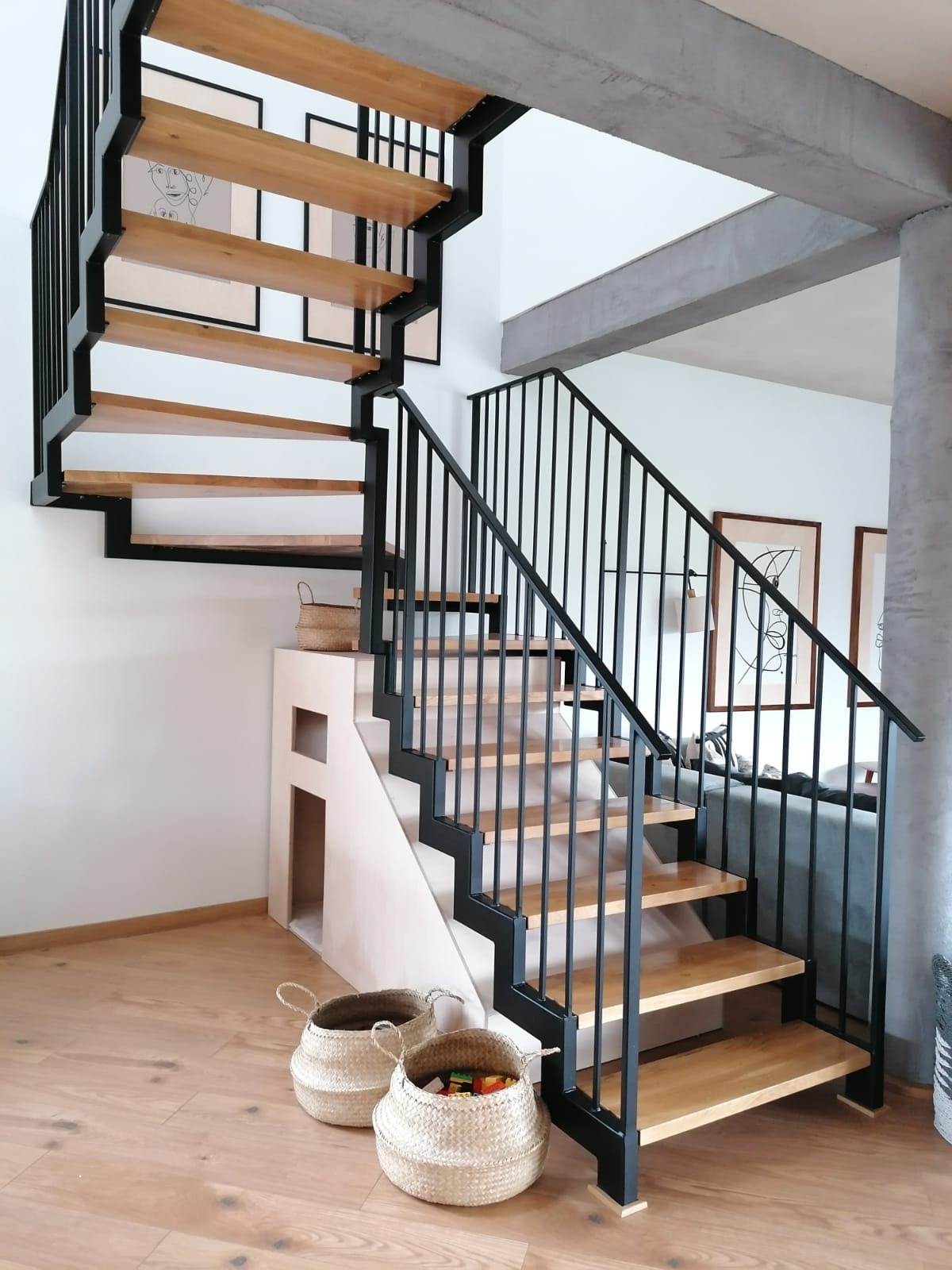 Nowoczesne schody metalowe z drewnianymi stopniami i czarną balustradą w jasnym wnętrzu. Widoczne kosze wiklinowe i minimalistyczne obrazy na ścianie.