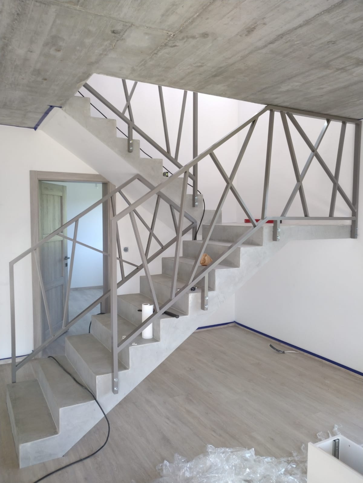 Betonowe schody z metalową, geometryczną balustradą w surowym wnętrzu. Widoczne zabezpieczenia malarskie, co sugeruje etap wykończeniowy.