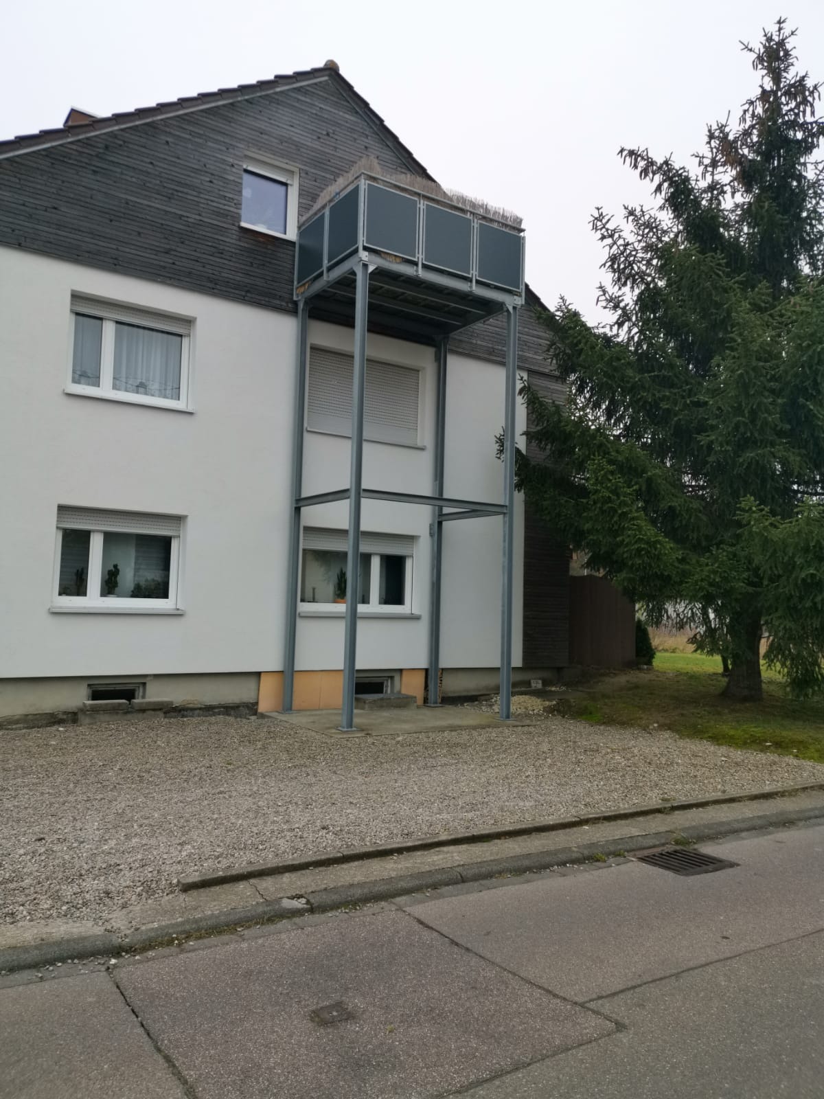 Metalowa konstrukcja balkonu zewnętrznego, przymocowana do ściany budynku. Szara kolorystyka, widok z poziomu ulicy, drzewo iglaste w tle.