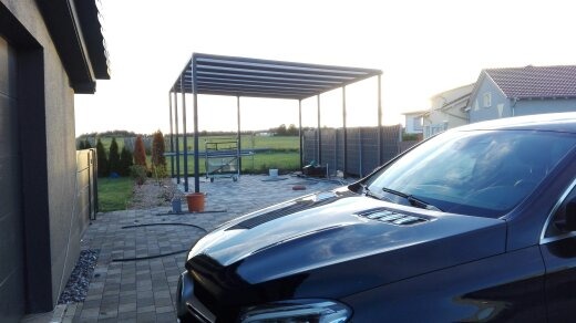 Nowoczesna, metalowa altana z zadaszeniem lamelowym na posesji, widoczna zza czarnego samochodu. W tle dom i ogrodzenie panelowe.