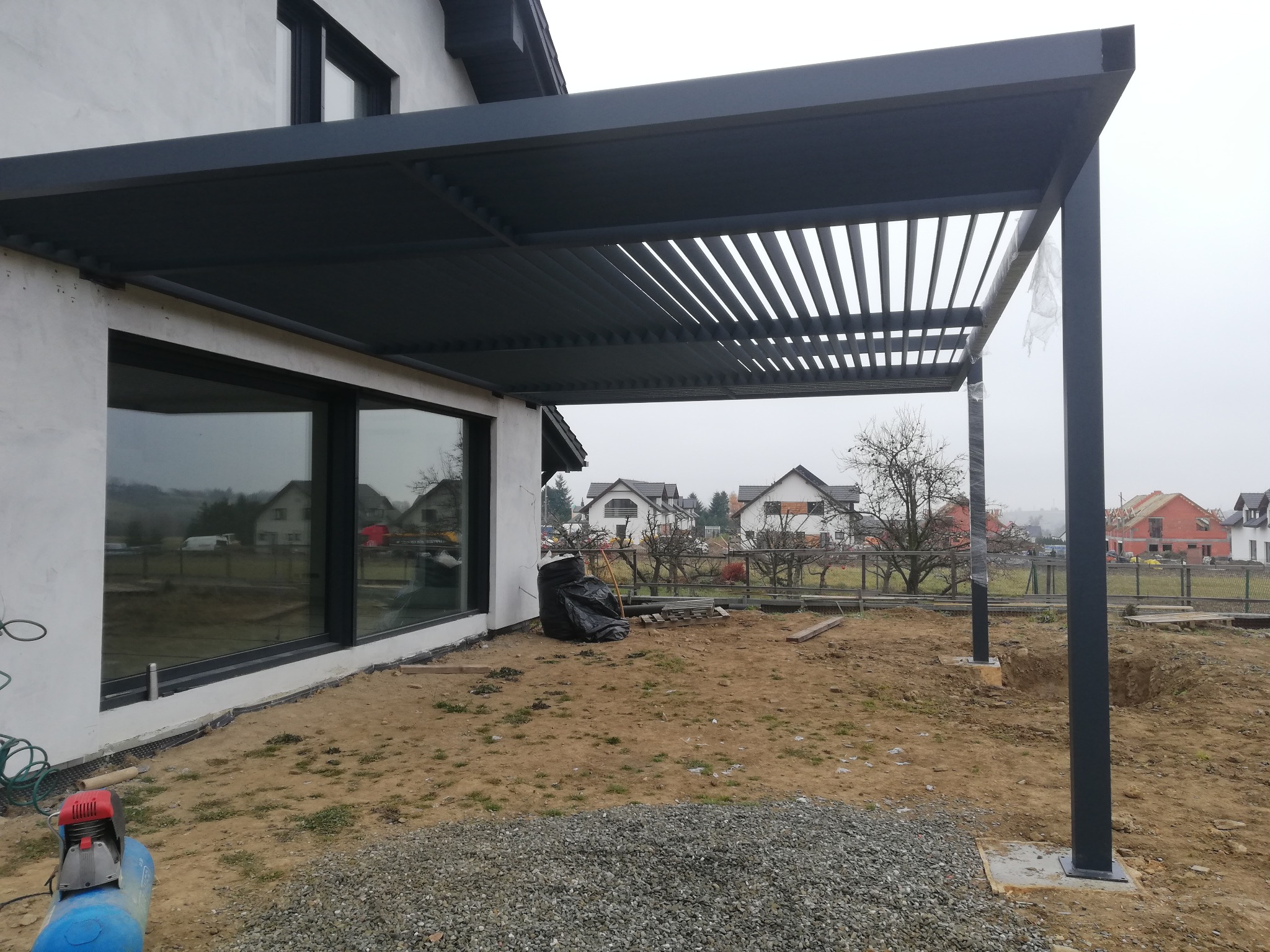 Nowoczesna, grafitowa altana z lamelowym dachem, przylegająca do budynku z dużymi oknami. Widoczne słupy i przygotowany teren pod dalsze prace.