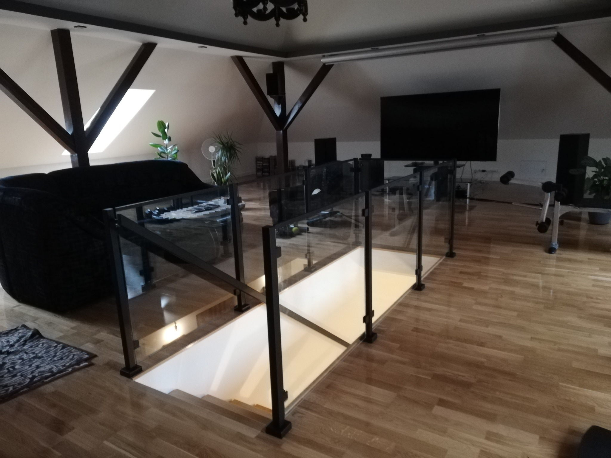 Szklana balustrada schodowa z czarnymi słupkami w nowoczesnym wnętrzu z drewnianą podłogą i widocznymi belkami stropowymi. Minimalistyczny design.