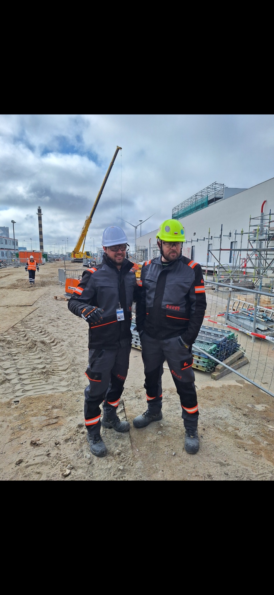 Dwóch pracowników w kaskach i kombinezonach roboczych na placu budowy z dźwigiem w tle. Widoczne rusztowania i elementy konstrukcyjne.