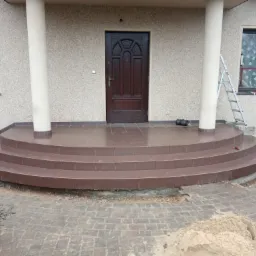 Nowe, brązowe, kafelkowe schody wejściowe do domu z kolumnami, częściowo wykończone, z widocznymi pozostałościami po pracach brukarskich i narzędziami.