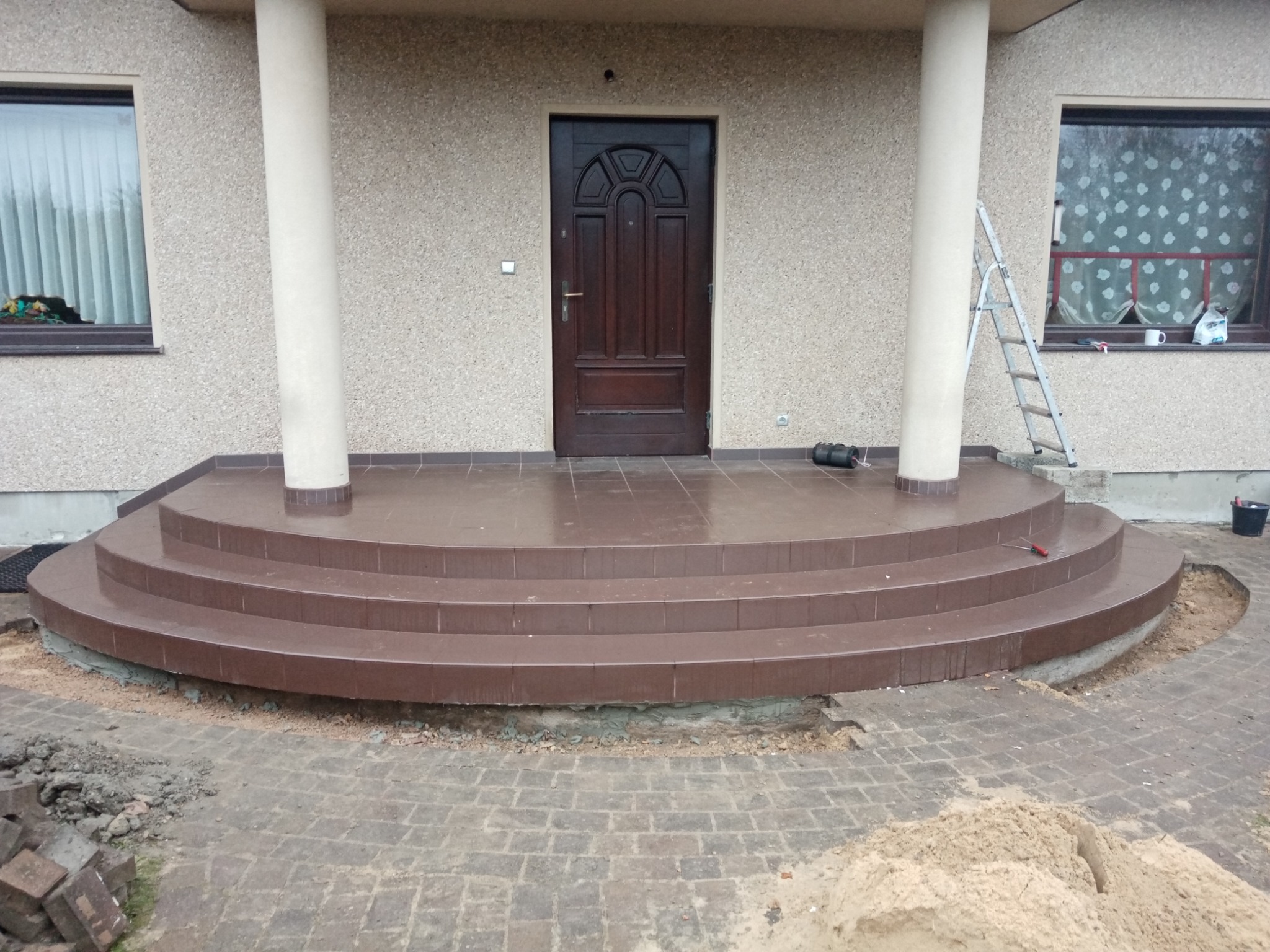 Nowe, brązowe, kafelkowe schody wejściowe do domu z kolumnami, częściowo wykończone, z widocznymi pozostałościami po pracach brukarskich i narzędziami.