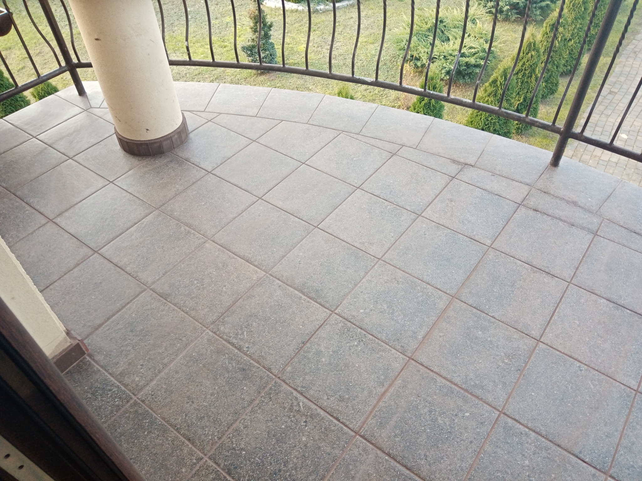 Wyłożony kwadratowymi płytkami balkon z metalową balustradą i widokiem na drzewa iglaste w ogrodzie.