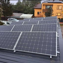Fotowoltaika Sunsate Wojciech Heidinger – Instalacja | Montaż | Panele Fotowoltaiczne | Magazyny Energii - Rząd paneli słonecznych zamontowanych na ciemnoszarym blaszanym dachu domu jednorodzinnego z pomarańczową elewacją w tle.
