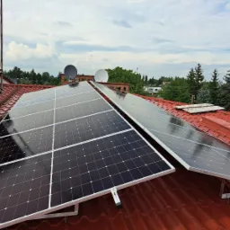 Panele słoneczne na czerwonym dachu z dachówki ceramicznej, widoczne anteny satelitarne, komin z cegły i fragmenty zieleni w tle, pochmurne niebo.