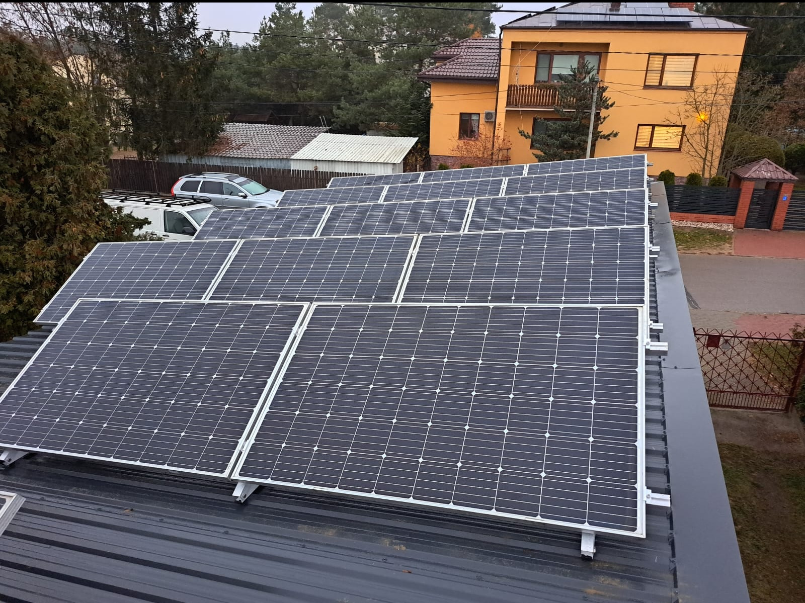 Rząd paneli słonecznych zamontowanych na ciemnoszarym blaszanym dachu domu jednorodzinnego z pomarańczową elewacją w tle.