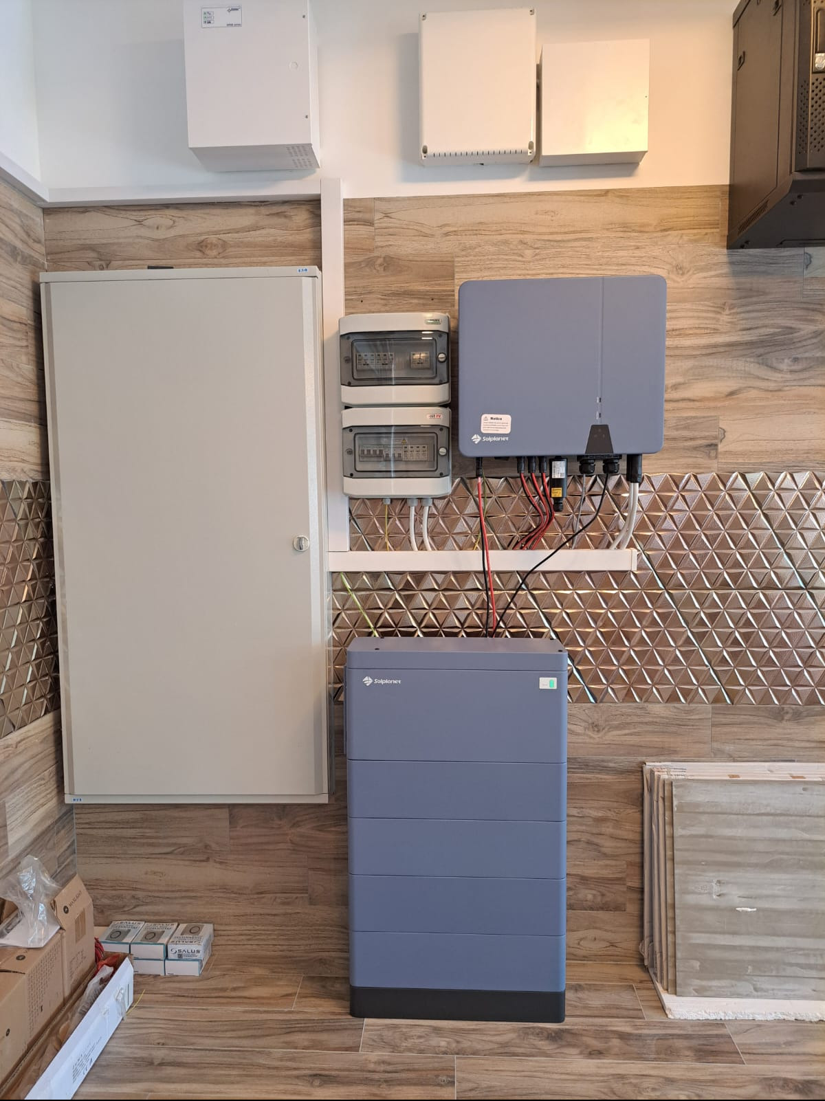 Szara bateria magazynu energii Solplanet zamontowana na ścianie wyłożonej płytkami imitującymi drewno, z widocznymi przewodami, obok skrzynka elektryczna i zabezpieczenia.