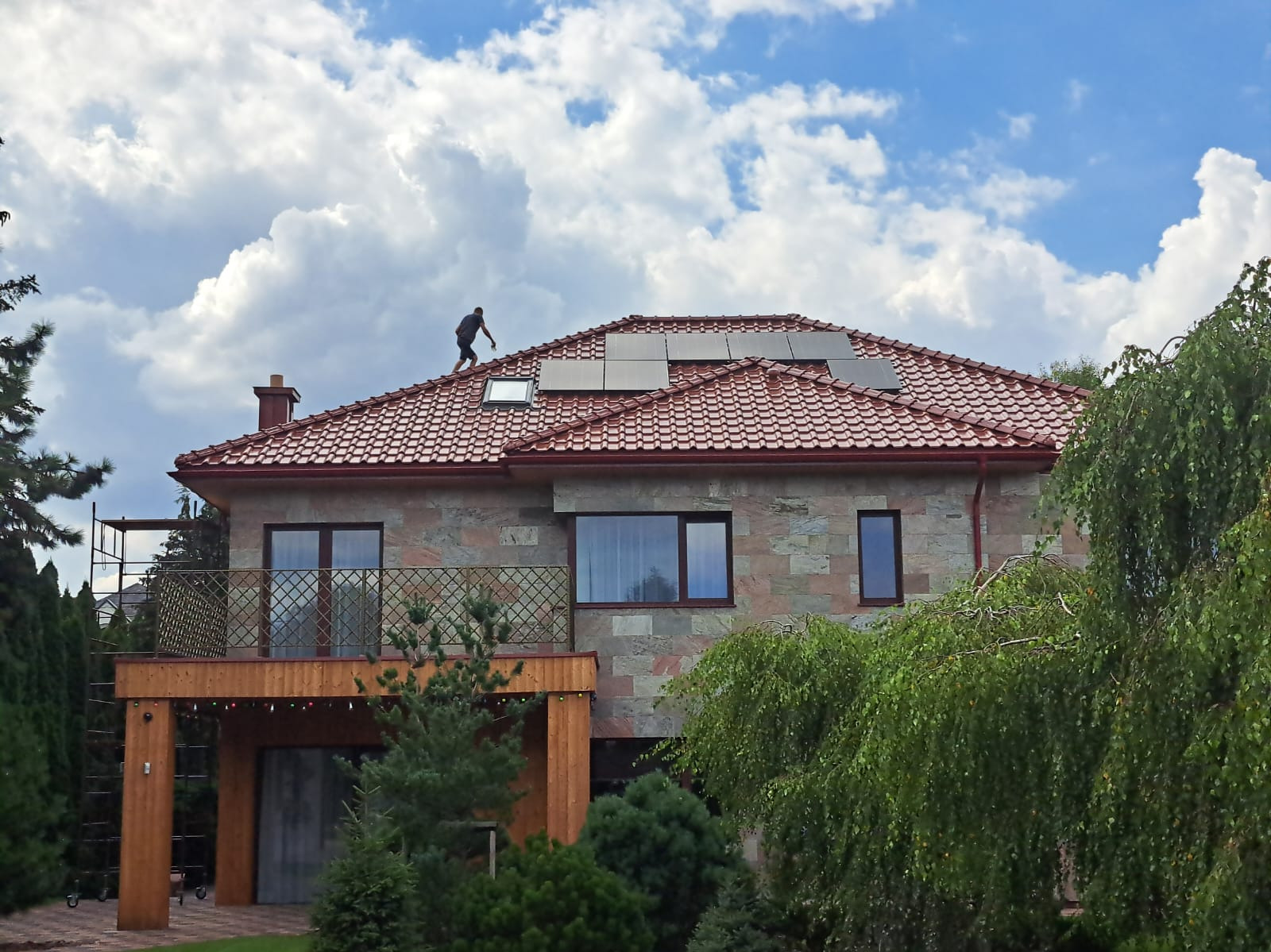 Montaż paneli słonecznych na dachu dwukondygnacyjnego domu z czerwoną dachówką, pracownik w ciemnej koszulce na dachu, błękitne niebo z chmurami w tle.