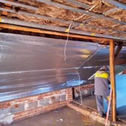 Prace izolacyjne poddasza w trakcie realizacji: widoczna wełna mineralna między krokwiami, folia aluminiowa oraz pracownik w odzieży roboczej przytwierdzający niebieską płytę izolacyjną...