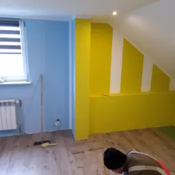 Instalacja paneli podłogowych w pokoju z pomalowanymi na niebiesko i żółto ścianami. Widoczny pracownik w trakcie układania paneli, szczotka i grzejnik.
