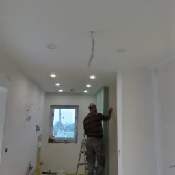 Pracownik na drabinie szpachluje ścianę z płyty gipsowo-kartonowej w trakcie remontu, widoczne okno i instalacja elektryczna sufitowa