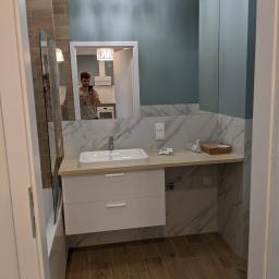 High Standard  - wykończenia wnętrz - Wykończona łazienka z umywalką nablatową na szafce z szufladami, marmurowe płytki na ścianach, drewnopodobna podłoga, widoczne akcesoria montażowe i karton na blacie, odbicie fotografa w lustrze.