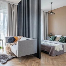 High Standard  - wykończenia wnętrz - Nowoczesne wnętrze z drewnianą podłogą w jodełkę, szarą sofą z dekoracyjnymi poduszkami, czarną ścianką działową z pionowych listew oddzielającą część sypialnianą z łóżkiem i beżową ścianą,...