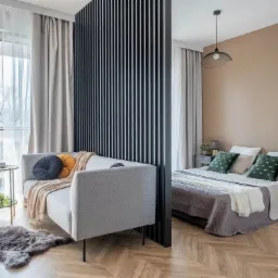 Nowoczesne wnętrze z drewnianą podłogą w jodełkę, szarą sofą z dekoracyjnymi poduszkami, czarną ścianką działową z pionowych listew oddzielającą część sypialnianą z łóżkiem i beżową ścianą,...