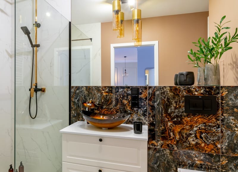 Nowoczesna łazienka z kabiną prysznicową typu walk-in, umywalką w kształcie misy na szafce z białymi frontami i okładziną ścienną z płytek imitujących marmur w odcieniach brązu i czerni, widoczne...