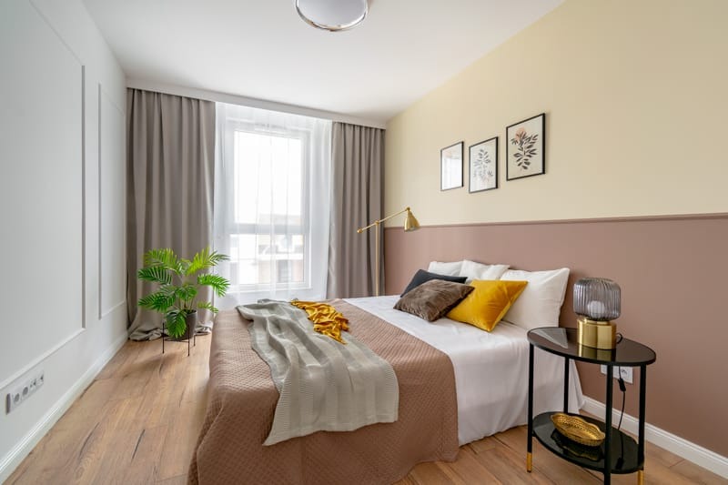 Jasna sypialnia z drewnianą podłogą, łóżkiem z brązową narzutą i żółtymi akcentami, ścianą podzieloną na kremową i różową część z trzema obrazkami, oraz małym czarnym stolikiem nocnym z lampką.