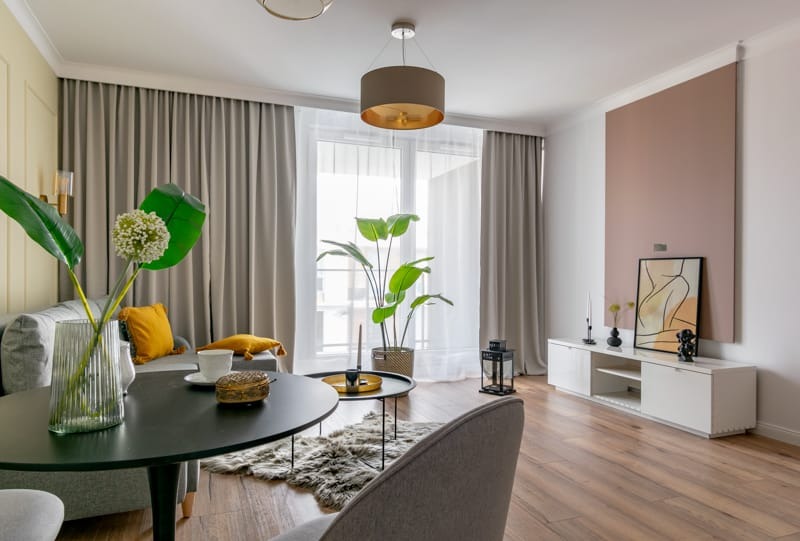 Jasne wnętrze salonu z drewnianą podłogą, szarymi zasłonami i minimalistycznymi meblami, akcentowane roślinami i dekoracjami w odcieniach złota i żółci.