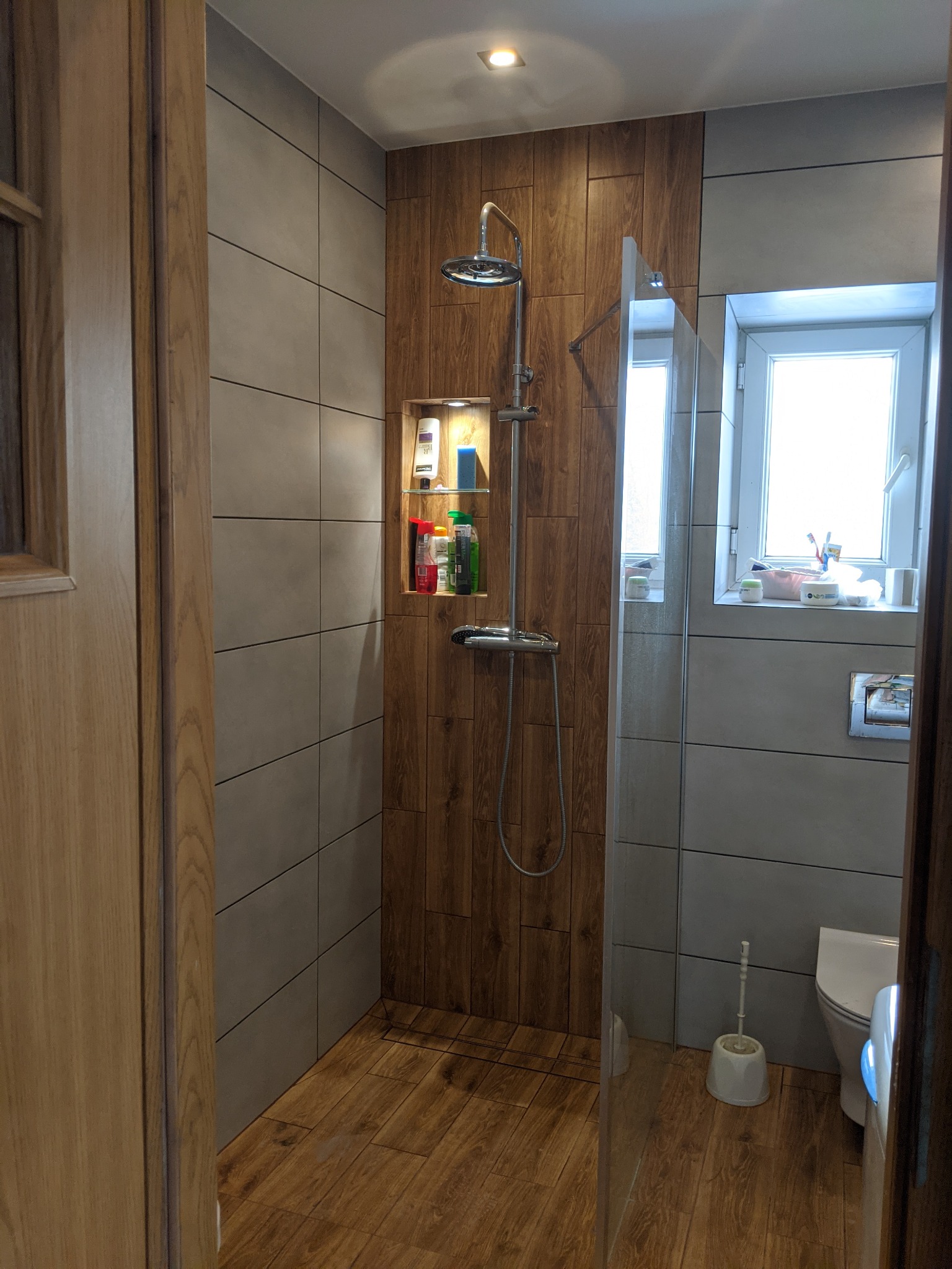 Nowoczesna łazienka z kabiną prysznicową typu walk-in, wykończona szarymi i drewnopodobnymi płytkami, z wnęką na kosmetyki podświetloną punktowym światłem.