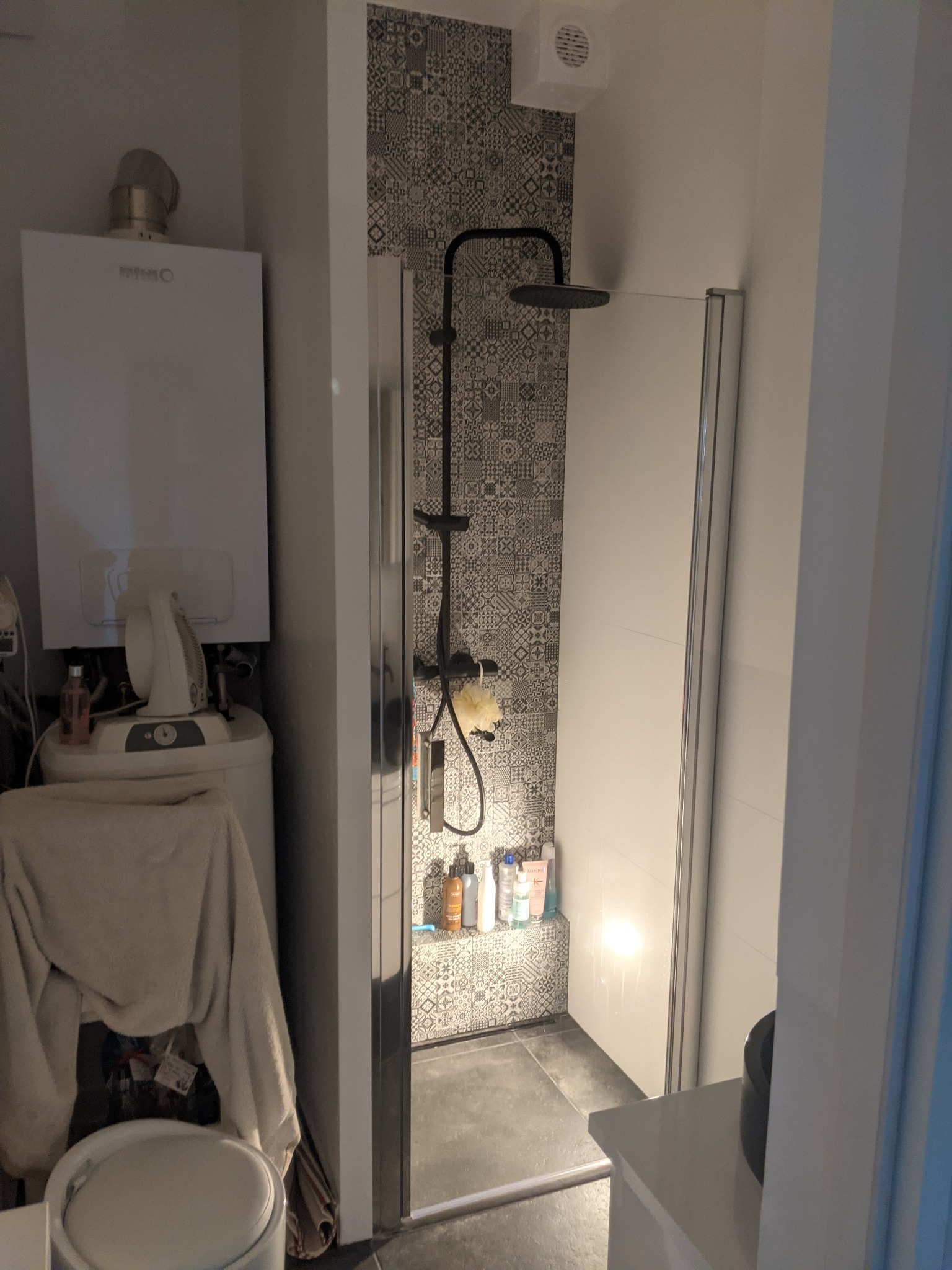 Wnętrze małej łazienki po remoncie: kabina prysznicowa z deszczownicą na tle ściany wyłożonej płytkami w geometryczne wzory, obok piec gazowy i pralka.