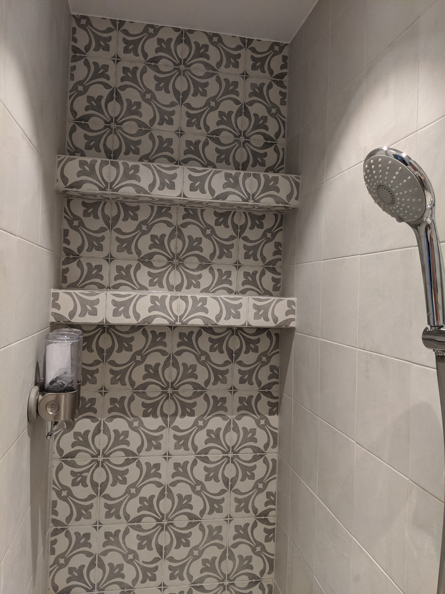 Wnęka prysznicowa wyłożona szarymi płytkami w kwiatowy wzór, z dwiema półkami z płytek i dozownikiem mydła w kolorze srebrnym.
