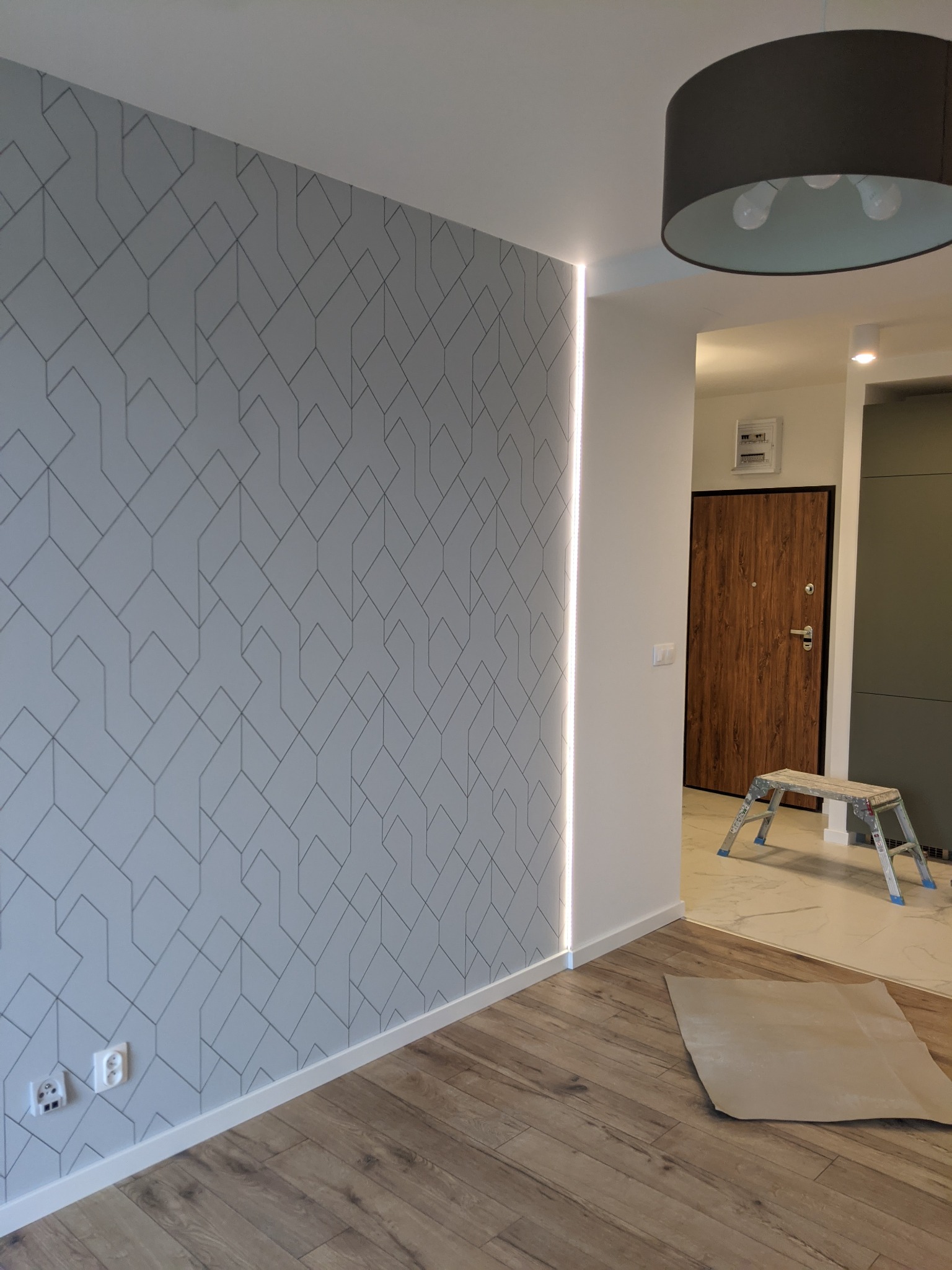 Szara ściana z geometrycznym wzorem i pionowym oświetleniem LED, drewniana podłoga, widoczny fragment sufitu z lampą, w tle drzwi i drabina malarska.