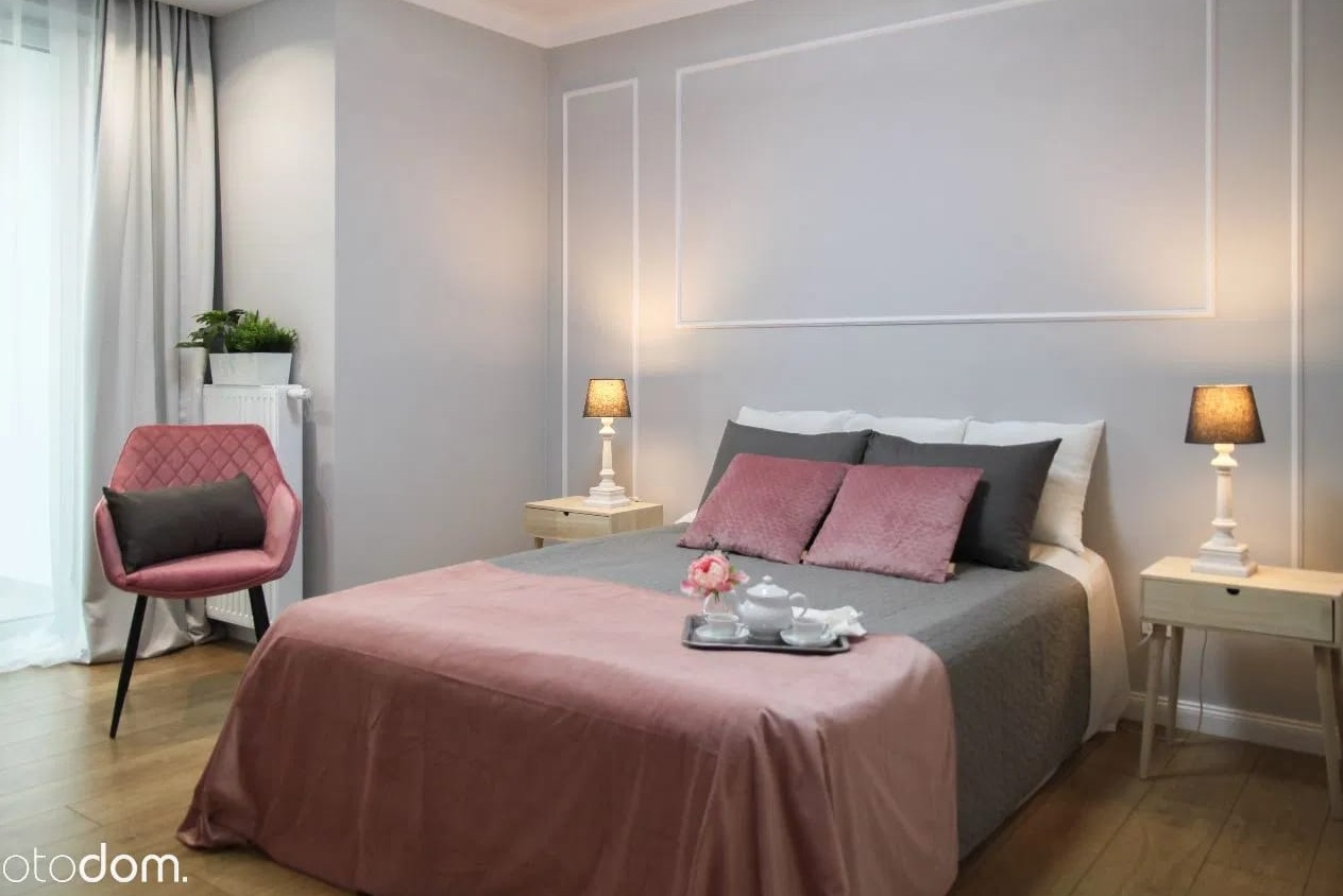 Jasnoszary pokój z łóżkiem, różowym pledem i poduszkami, stolikami nocnymi z lampkami, różowym krzesłem i sztukaterią na ścianach.