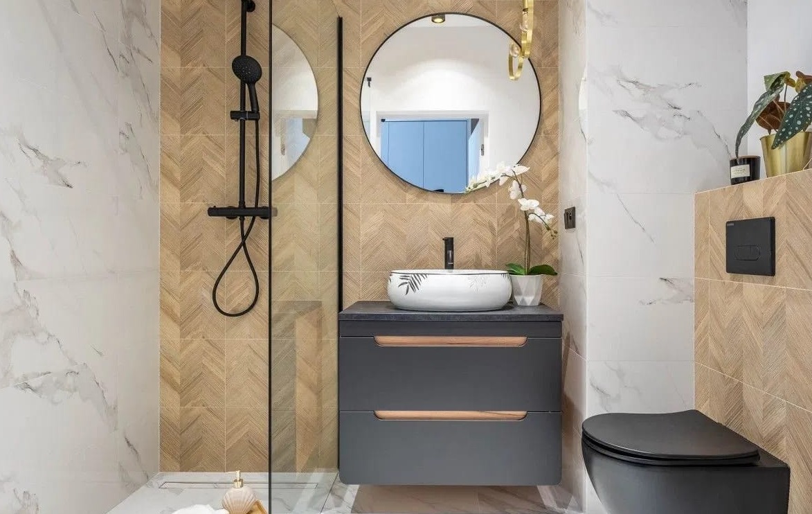 Nowoczesna łazienka z kabiną prysznicową typu walk-in, umywalką nablatową z motywem roślinnym, okrągłym lustrem i płytkami imitującymi drewno ułożonymi w jodełkę.
