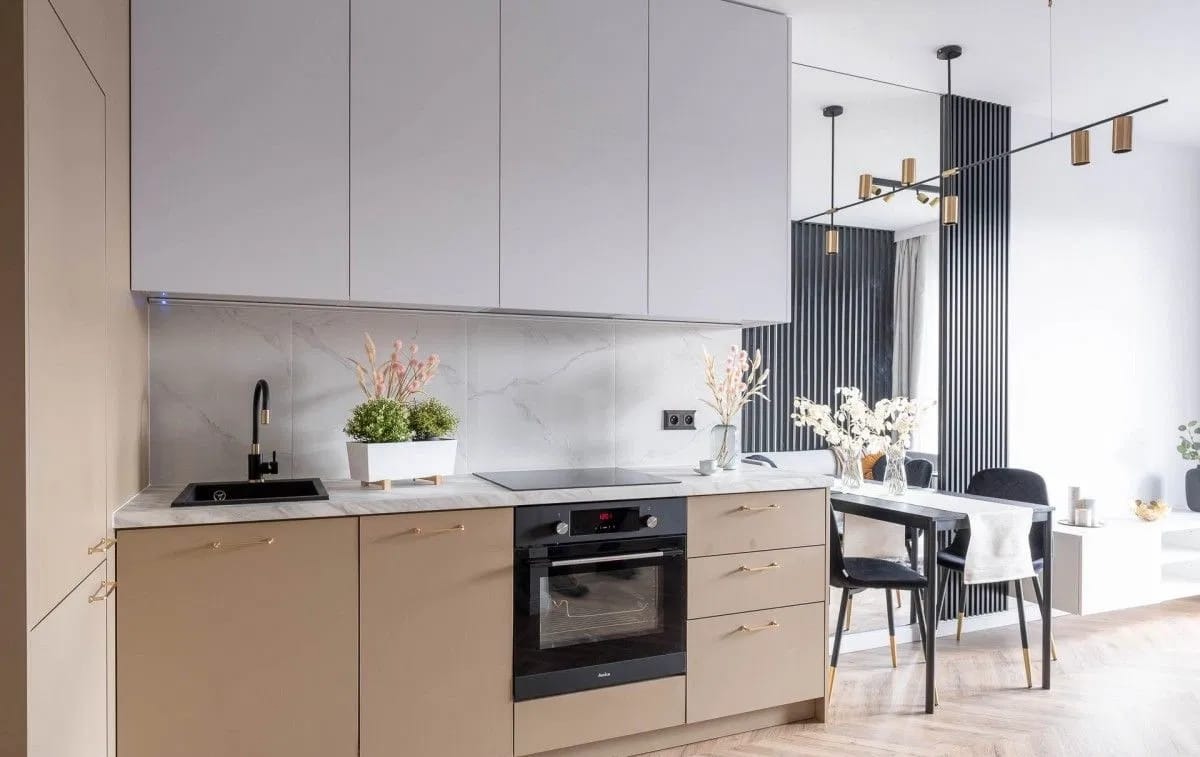 Nowoczesna kuchnia w odcieniach beżu i szarości z marmurowym blatem, czarną armaturą i złotymi uchwytami, uzupełniona o elementy dekoracyjne takie jak kwiaty w wazonie i doniczce.