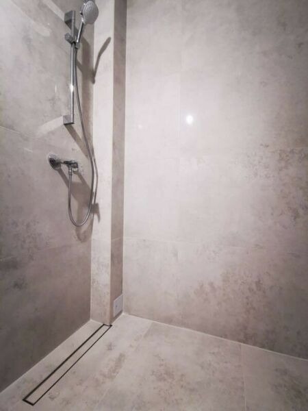 Wykończona kabina prysznicowa z odpływem liniowym, wyłożona płytkami imitującymi kamień w odcieniach szarości i beżu, z zamontowaną baterią i deszczownicą.