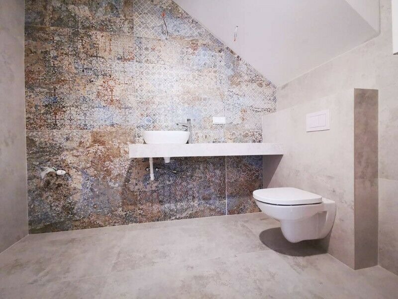 Wykończona łazienka z umywalką nablatową na białym blacie i podwieszaną toaletą, ściana wyłożona płytkami o wzorze imitującym stare freski w odcieniach brązu, błękitu i szarości.