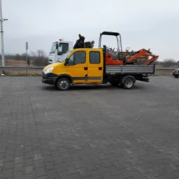 Żółty samochód dostawczy z pomarańczową mini koparką na platformie, stojący na parkingu z kostki brukowej, w tle biała ciężarówka i zalesiony teren w pochmurny dzień.