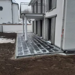 Nowoczesny taras z szarej kostki brukowej ułożonej przy domu z szarymi okiennicami, widok na kościół w tle, częściowo pokryty śniegiem.