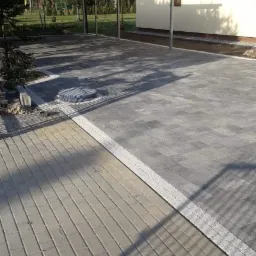 Ułożona szara kostka brukowa przed domem z wiatą, widoczny fragment chodnika z jaśniejszej kostki i studzienka kanalizacyjna.